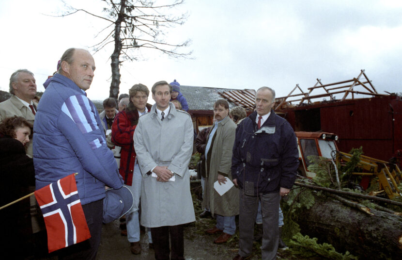 Rovde, Sunnmøre 19920108: Kong Harald besøker orkanrammede på Sunnmøre. Kong Harald (t.v.) blir her orientert av ordfører Leif Helge Kongshaug (midten) under et besøk på gårdsbruket til Aarskog i Rovde kommune. Skadene etter orkanen kommer opp i 1,3 mrd. kroner.Norsk flagg. Kongen i blå boblejakke. (Foto: Bjørn Sigurdsøn/NTB.)